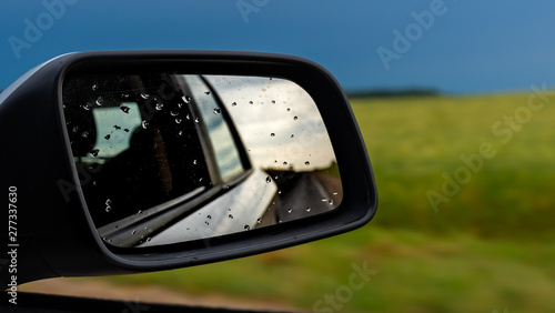 raindrops on side rear-view mirror on a car in a raining day. drops of rain on car window. blue sky, sun light, green field over rainy wet road. driving in bad weather.