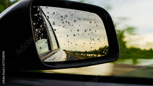 raindrops on side rear-view mirror on a car in a raining day. drops of rain on car window. sunset, sun light, green field over rainy wet road. driving in bad weather. © Natallia