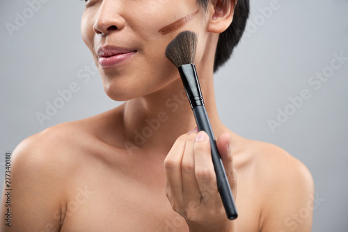 Close-up of young beautiful woman applying foundation cream with brush and correcting her cheek