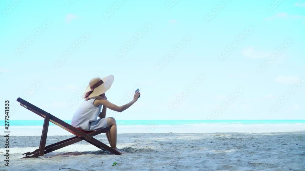 4K Young adult asian woman tourist in summer dress sitting on beach chair sun bed for relax sunbathing on island sea beach in summer holiday vacation travel using smartphone for selfie or photography 4K Young adult asian woman tourist in summer dress sitting on beach chair sun bed for relax sunbathing on island sea beach in summer holiday vacation travel using smartphone for selfie or photography