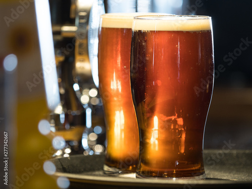 Two pints of dark coloured real ale stand on a tray in pint glasses with frothy heads back lit on a bar top. - Image