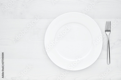 Empty white round ceramic plate and fork on white wooden table with copy space