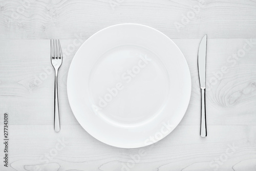 Empty white round ceramic plate, fork and knife on white wooden table with copy space