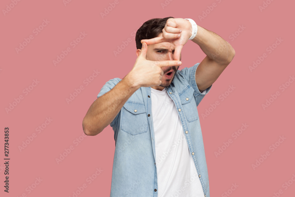 Portrait of attentive handsome bearded young man in blue casual style shirt standing crop composition focus hand gesture and looking at camera . indoor studio shot, isolated on pink background.