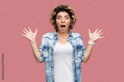 Portrait of surprised young woman with curly hairstyle in casual blue shirt standing with big eyes, open mouth and looking at camera with raised arms. indoor studio shot, isolated on pink background.