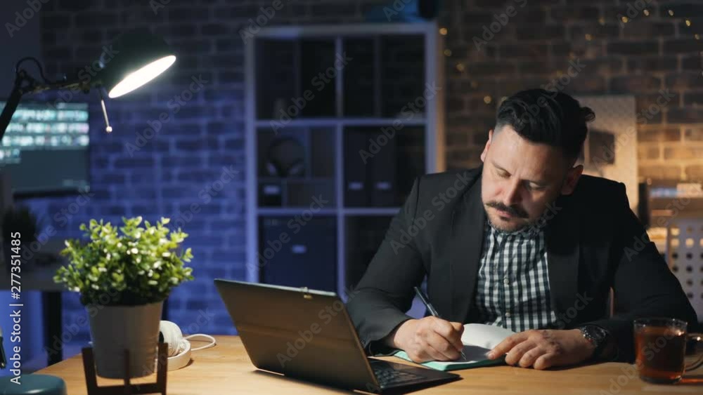 Handsome businessman is working with laptop at night in cozy office writing in notebook concentrated on activity. People, modern devices and business concept. Handsome businessman is working with laptop at night in cozy office writing in notebook concentrated on activity. People, modern devices and business concept.