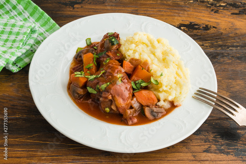 Coq au Vin, traditional French recipe of chicken braised in red wine with bacon, carrot and mushrooms. Served with mashed potatoes on white plate.