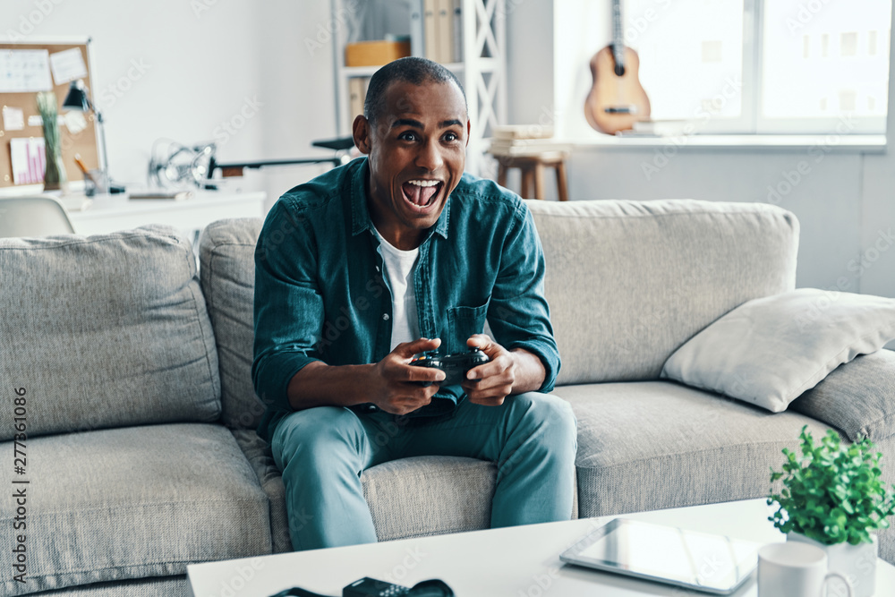 So much fun! Handsome young African man playing video game and smiling while sitting indoors So much fun! Handsome young African man playing video game and smiling while sitting indoors