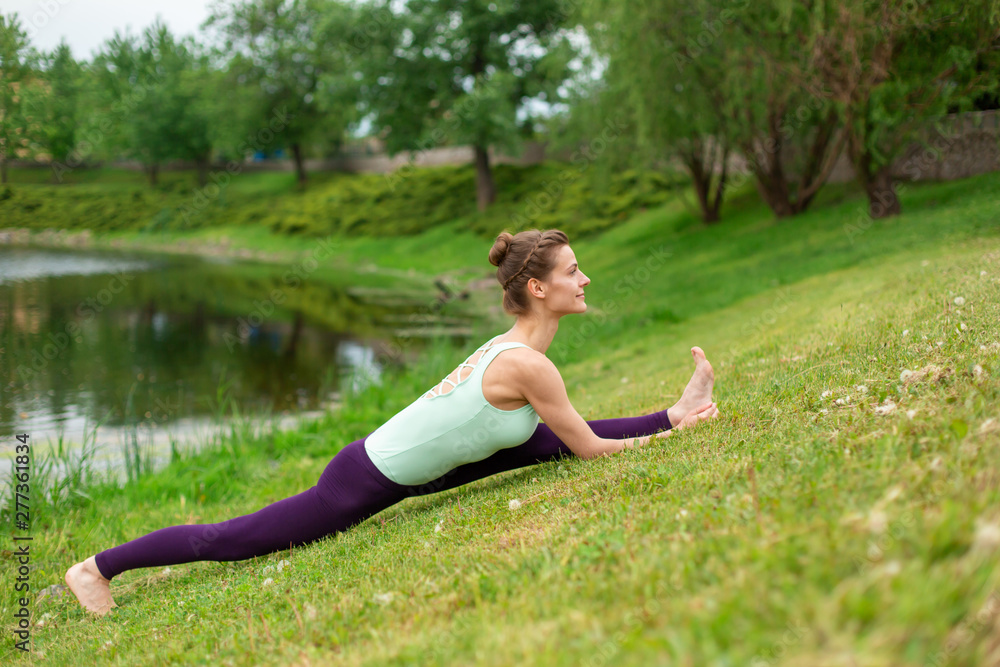 Slim young brunette girl yogi does difficult yoga exercises on the green grass on the background of water