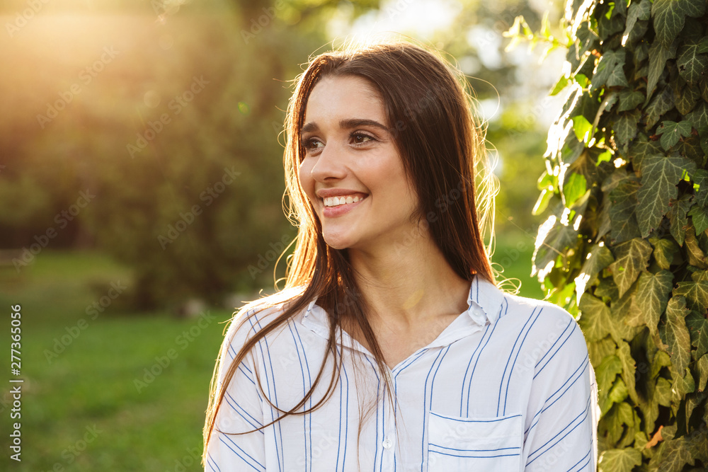 Photo of pretty young woman wearing striped shirt smiling and walking in green park Photo of pretty young woman wearing striped shirt smiling and walking in green park
