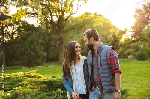 Image of laughing couple man and woman looking at each other while walking together in green park