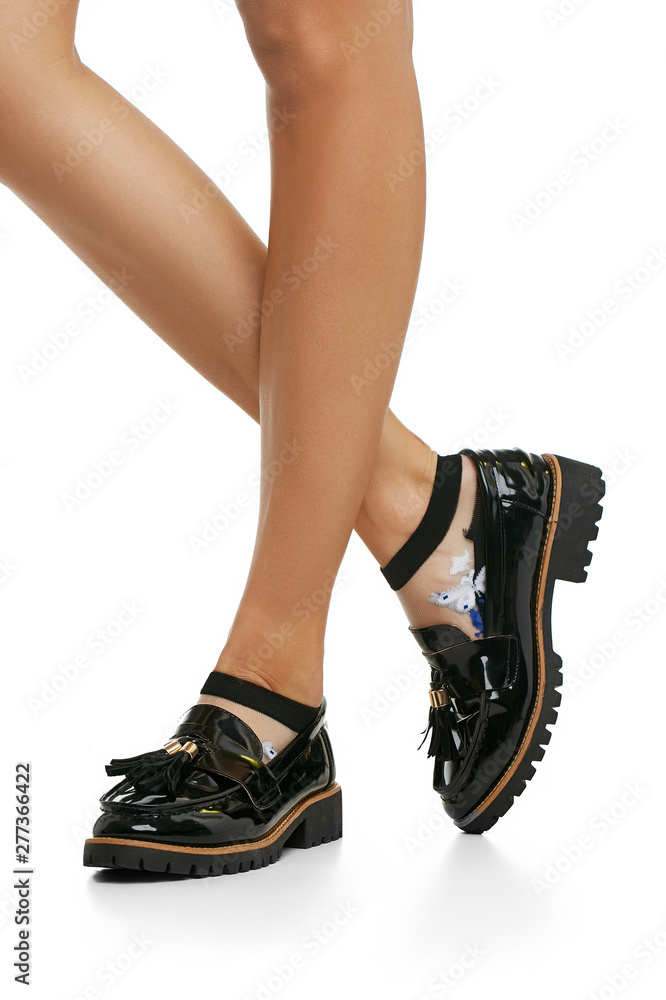 Cropped side view of lady's legs wearing nylon low-cut socks with embroidery in view of gray butterflies. The woman is wearing patent leather shoes with ribbed soles and tassels on white background.