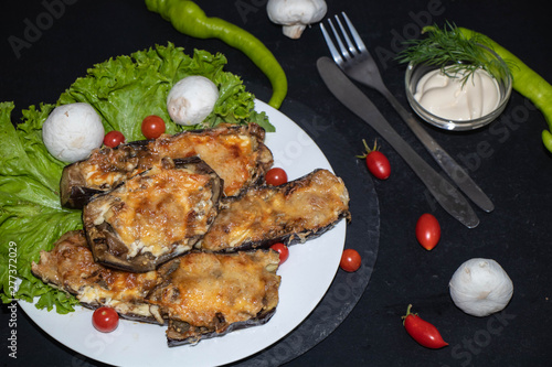 Eggplant stuffed with mushrooms and onions, rubbed with cheese, baked in the oven next to cherry tomatoes, lettuce. In the bowl sauce. All on a black background.