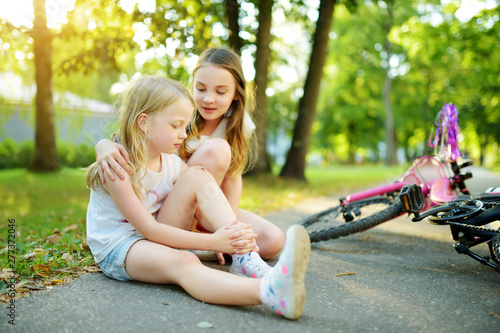 Adorable girl comforting her little sister after she fell off her bike at summer park. Child getting hurt while riding a bicycle.