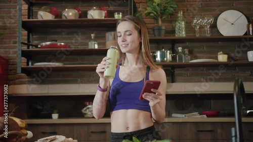 Cheerful young stylish woman in sportswear standing in modern kitchen and actively surfing the internet via phone, then takes a sip of organic smoothie and looks straight to camera, smiling cutely