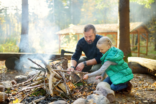Cute young girl learning to start a bonfire. Father teaching her daughter to make a fire. Child having fun at camp fire. Camping with kids in fall forest.