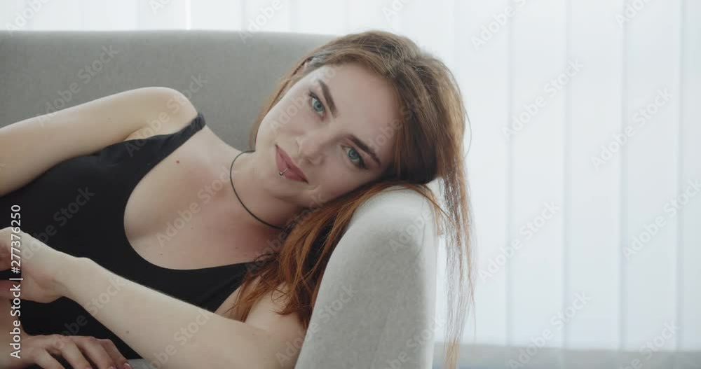 Closeup shoot of young seductive caucasian female in black bodysuit laying on the couch looking at camera and smiling indoors Closeup shoot of young seductive caucasian female in black bodysuit laying on the couch looking at camera and smiling indoors