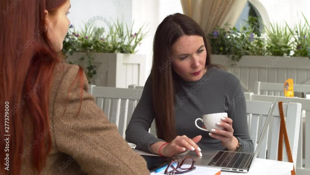 Two caucasian women watch the examples of necessary documents at the laptop screen sitting at the cafe slow motion Two caucasian women watch the examples of necessary documents at the laptop screen sitting at the cafe slow motion