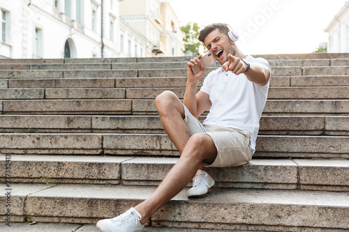 Portrait of joyful young man wearing headphones holding smartphone and singing outdoors