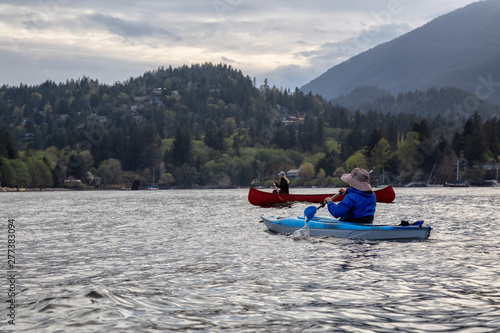 Adventurous friends on a red canoe and kayak are paddling in the Howe Sound during a cloudy sunset. Taken near Bowen Island, West of Vancouver, BC, Canada.