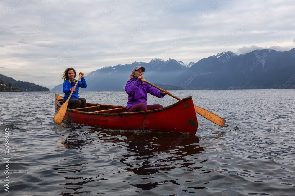 Couple adventurous female friends on a red canoe are paddling in the Howe Sound during a cloudy sunset. Taken near Bowen Island, West of Vancouver, BC, Canada.
