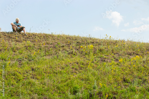 A young man sits on top of a hill and looks into the phone. A tourist is resting on a hike, checked with a map in the navigator. man sits on a hill wearing a hat with a smartphone in his hands