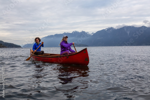 Couple adventurous female friends on a red canoe are paddling in the Howe Sound during a cloudy sunset. Taken near Bowen Island  West of Vancouver  BC  Canada.