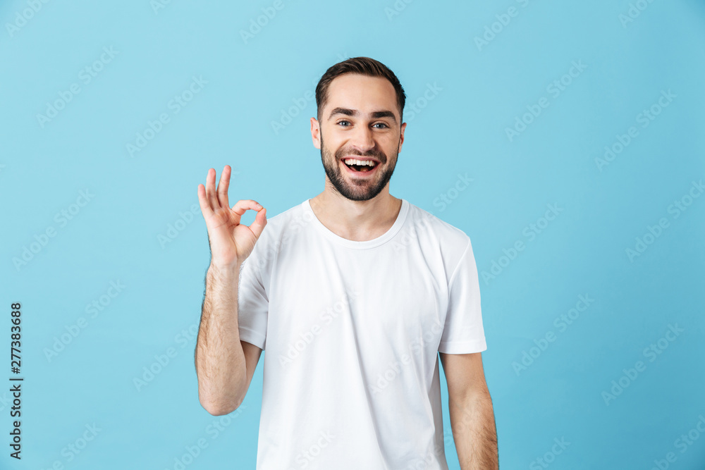 Smiling young happy bearded man posing isolated over blue wall background showing okay gesture. Summer concept. Smiling young happy bearded man posing isolated over blue wall background showing okay gesture. Summer concept.