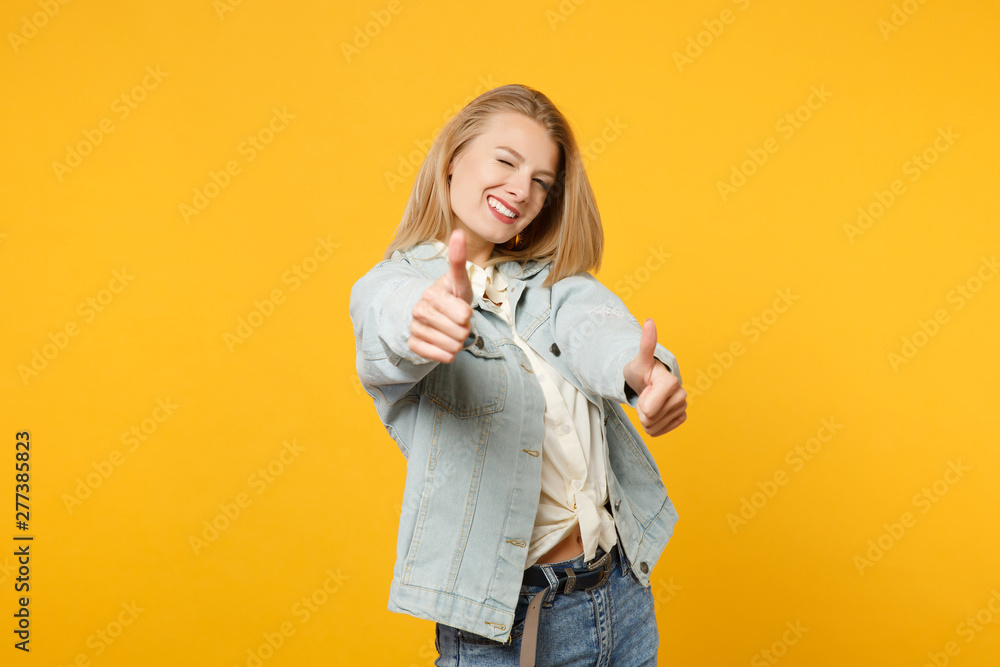 Fototapeta premium Portrait of cheerful young woman in denim casual clothes looking camera and showing thumbs up isolated on bright yellow orange background wall in studio. People lifestyle concept. Mock up copy space.