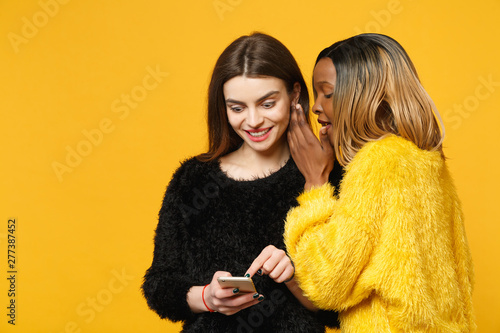 фотографія Two young women friends european and african american in black yellow clothes standing posing isolated on bright orange wall background, studio portrait