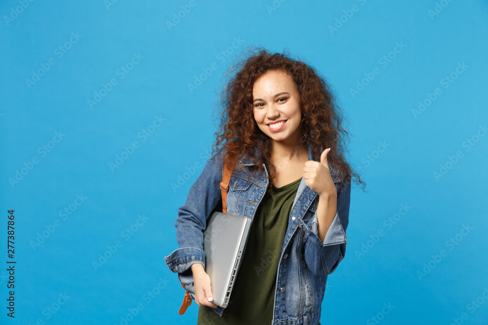 Obraz premium Young african american girl teen student in denim clothes, backpack hold pc isolated on blue wall background studio portrait. Education in high school university college concept. Mock up copy space.