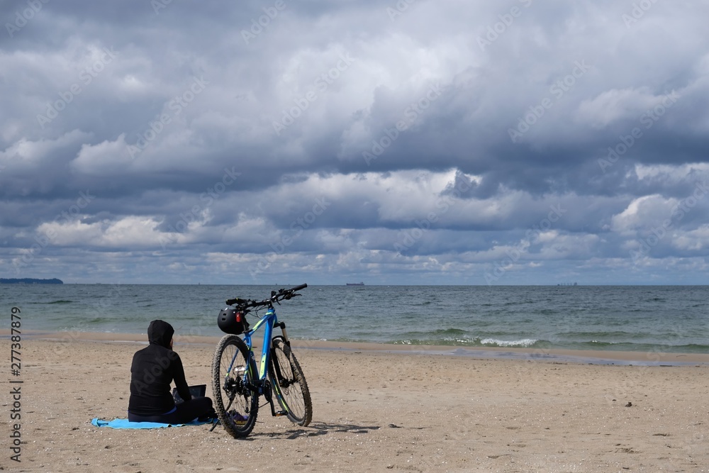Poland, Gdansk, Baltic Sea - women in black cloth sitting on the beach with bike in cloudy day with amazing dramatic clouds
