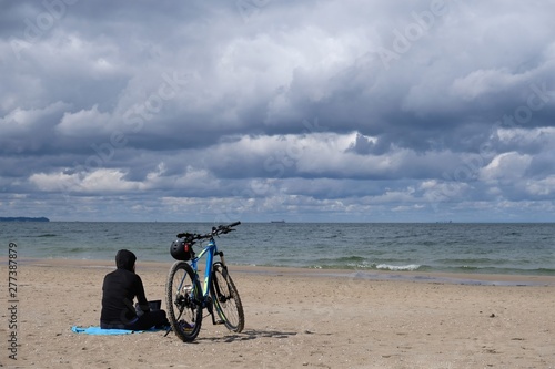 Poland, Gdansk, Baltic Sea - women in black cloth sitting on the beach with bike in cloudy day with amazing dramatic clouds