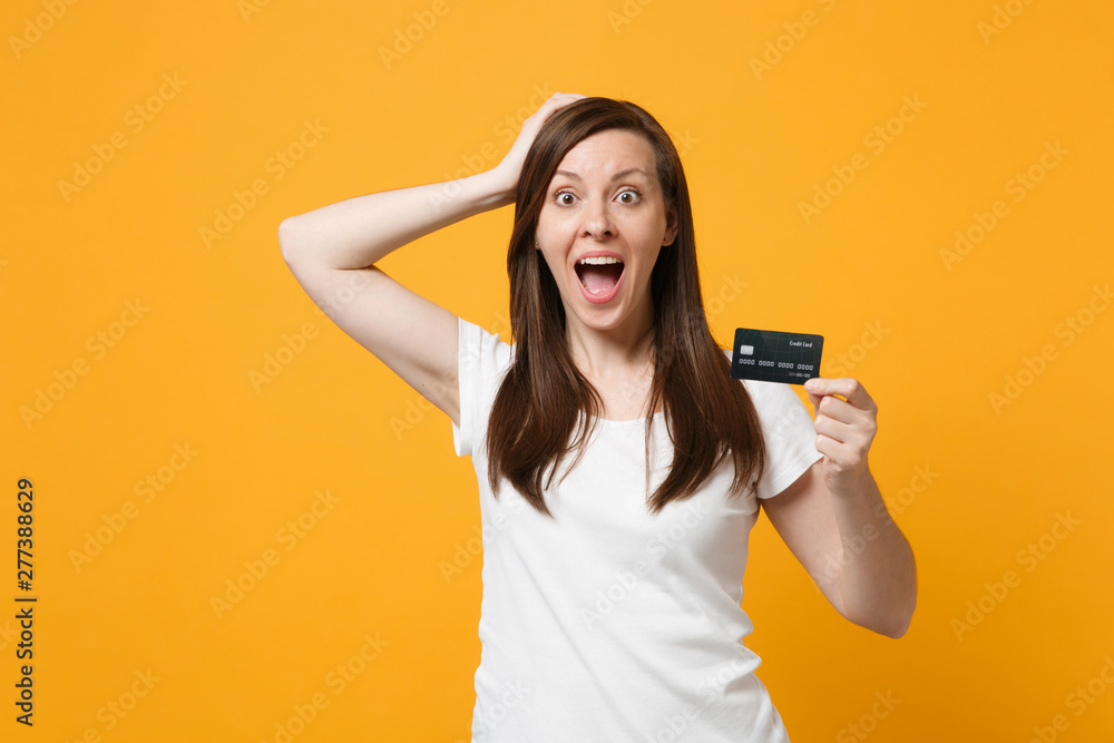 Portrait of surprised young woman in casual clothes keeping mouth open, putting hand on head, hold credit bank card isolated on yellow orange background. People lifestyle concept. Mock up copy space. - obrazy, fototapety, plakaty 