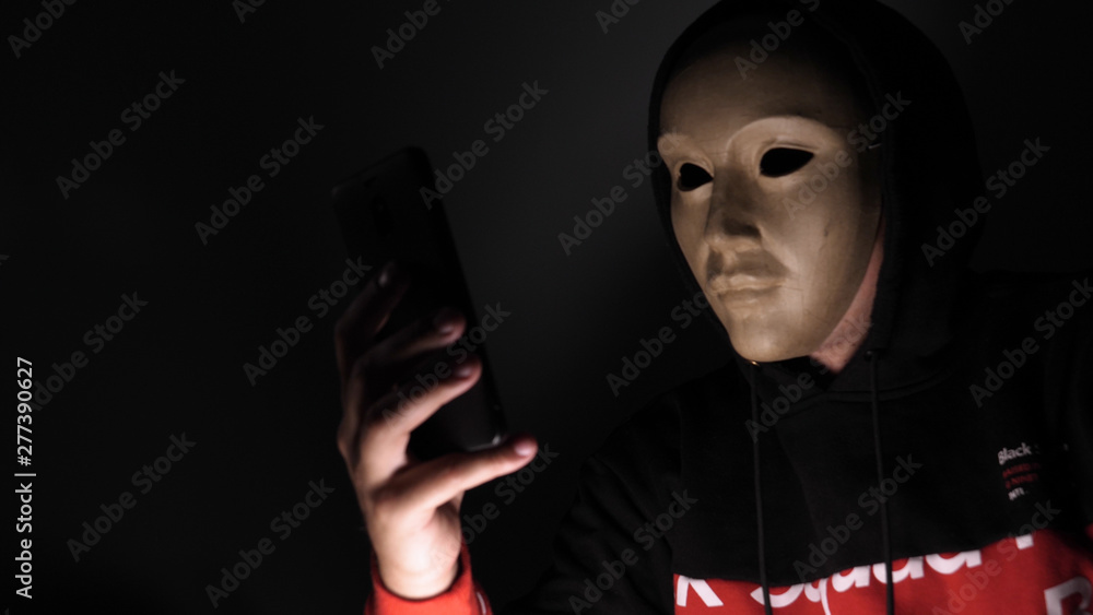 Male hacker hidden face with the mask accessing to personal information on laptop (phone) in the dark. Technology, cyber crime concept. Male hacker hidden face with the mask accessing to personal information on laptop (phone) in the dark. Technology, cyber crime concept.