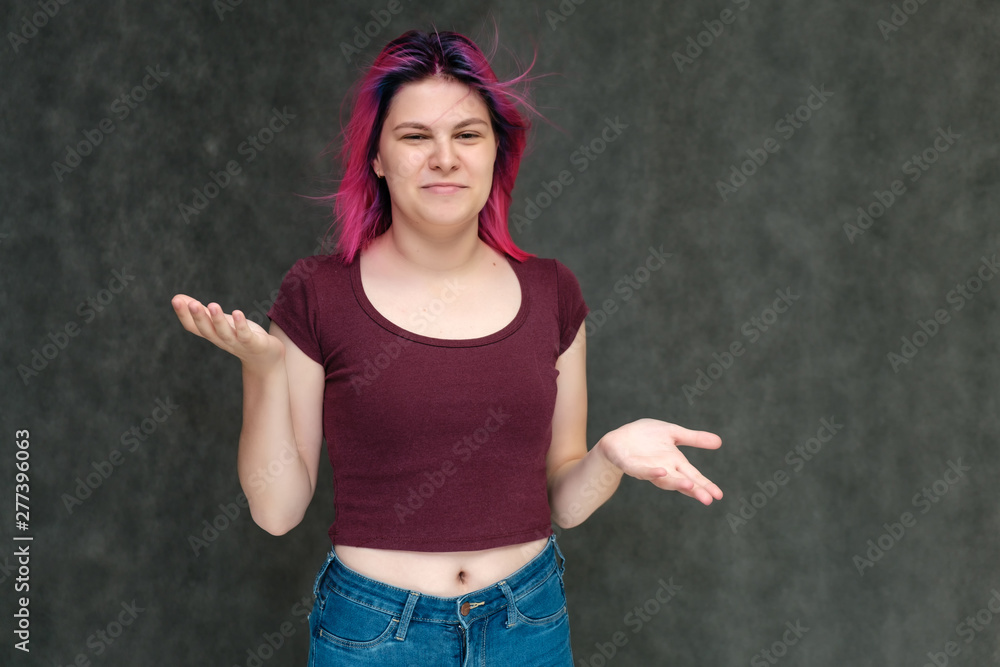 Portrait to the waist of a young pretty teen girl in a burgundy T-shirt and jeans with beautiful purple hair on a gray background in the studio. Talking, smiling, showing hands with emotions.