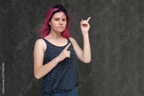 Portrait to the waist of a young pretty girl teenager in a T-shirt with beautiful purple hair on a gray background in the studio. Talking, smiling, showing hands with emotions.