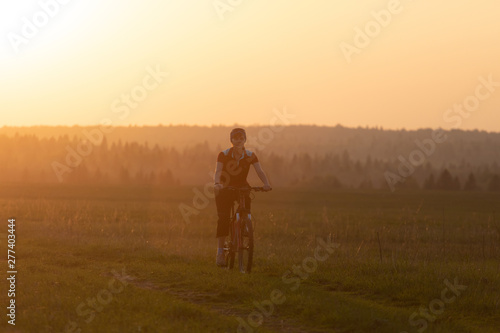 A girl rides a bicycle on the road against the backdrop of a bright sunset and yellow sun © Sergei Malkov