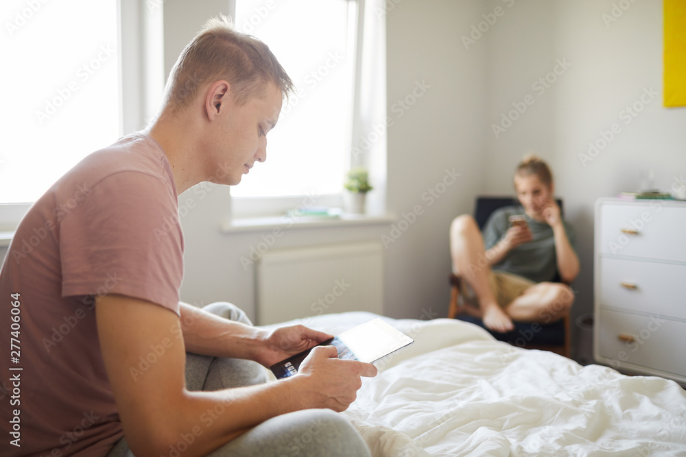 Fototapeta premium Contemporary guy with mobile gadget sitting on bed while scrolling in tablet with his wife on background