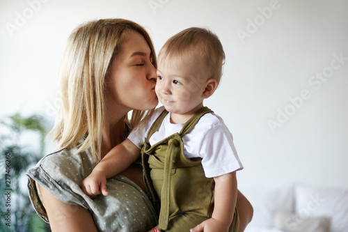 Indoor shot of attractive young European mother with blonde hair holding tight her cute chubby ten month old baby son, kissing him on cheek. Stylish mom bodning with adorable sweet infant child photo