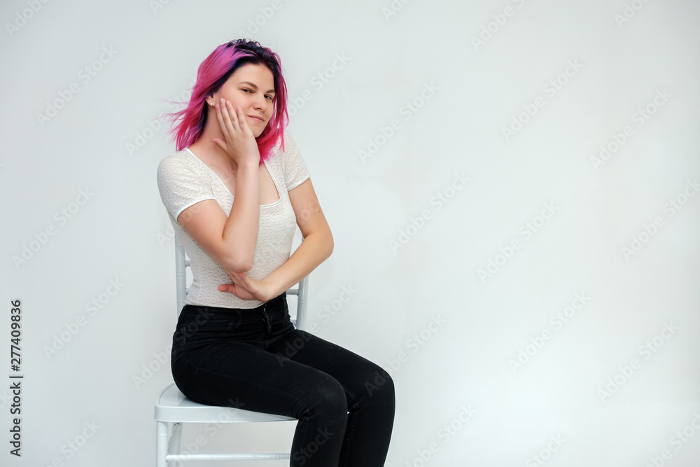 Portrait below the knee of a young beautiful girl teenager in a white T-shirt and black jeans with beautiful purple hair sitting on a white background in the studio. Says, shows hands with emotions. Portrait below the knee of a young beautiful girl teenager in a white T-shirt and black jeans with beautiful purple hair sitting on a white background in the studio. Says, shows hands with emotions.