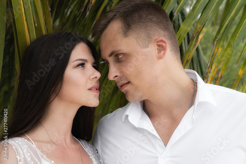 Newlyweds pierce each other's looks. A couple is standing under the leaves of a palm tree.