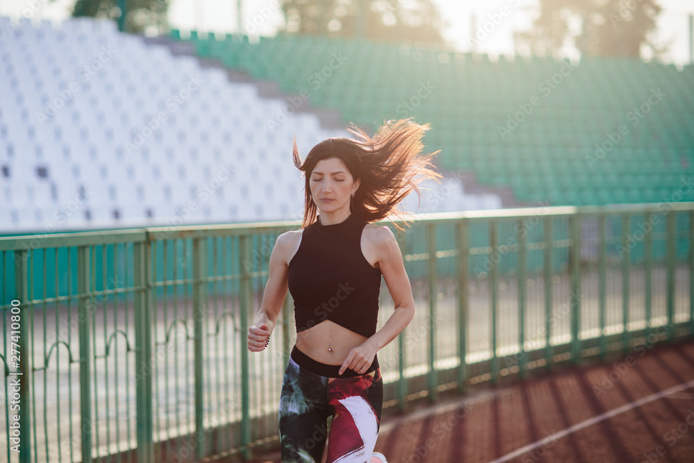 Sport. Athletic young brunette woman in pink sneakers, leggings and top run on running track stadium at sunset. her hair is developing. Concept run. concept of a healthy lifestyle.