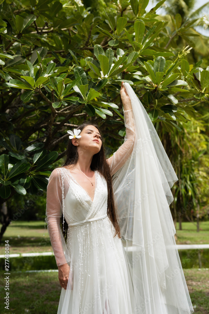 A bride in a white dress with an exotic flower in her hair is standing under a flowering tropical tree.