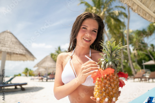 A brunette in a white bathing suit sits on a lounger with a Pina Colada cocktail in a pineapple