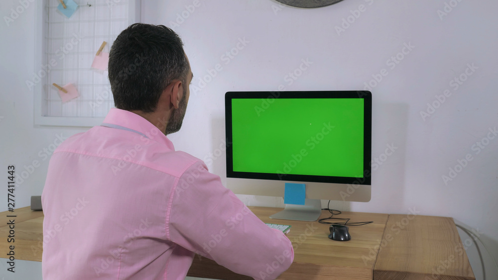 Happy smiling businessman turns and looking at the camera showing thumb signal thumbs-up. Handsome man sitting at the working place with wooden desk computer and green display Happy smiling businessman turns and looking at the camera showing thumb signal thumbs-up. Handsome man sitting at the working place with wooden desk computer and green display