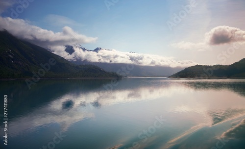 wispy clouds onto of snowy mountain peaks, with reflection in teal glacier waters in Alaska. 