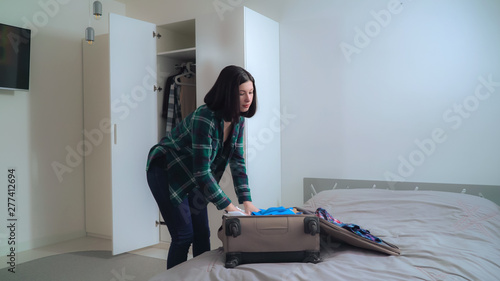 Young woman packing suitcase at home. Happy female pack up clothes in travel or business trip. Brunette choose what she want dress in vacation.