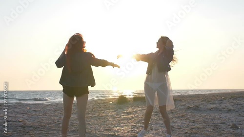 sunset rays over the sea illuminate the dance on the beach of two young sexy women dressed in boho style