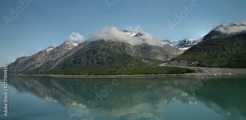 wispy clouds onto of snowy mountain peaks, with reflection in teal glacier waters in Alaska. 
